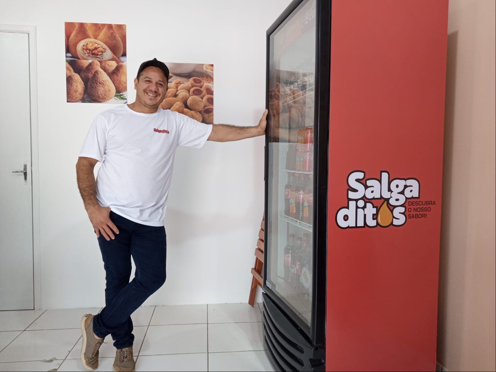 Salgaditos Um homem vestindo uma camiseta branca e calças escuras está de pé com uma mão apoiada em uma máquina de venda automática vermelha. A máquina tem o texto "Salgaditos: Descubra o Melhor Sabor" escrito nela. Há dois cartazes na parede atrás dele, ambos exibindo imagens de salgados. O chão é de azulejos e há uma porta fechada ao fundo.