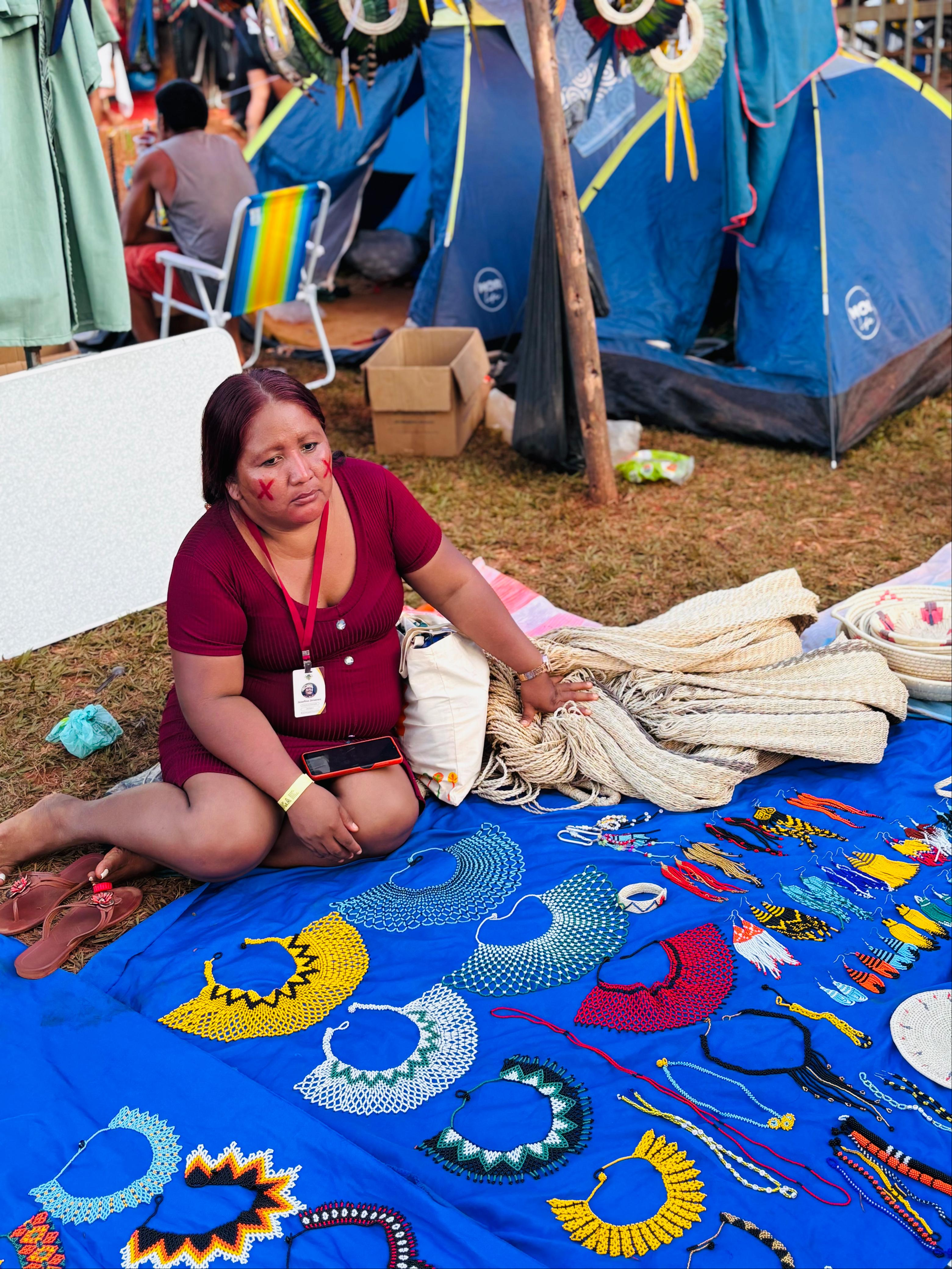 Indígena Warao expõe artesanatos no Acampamento Terra Livre 2025 Mulher indígena da etnia Warao está sentada em frente a uma barraca de camping, exibindo colares artesanais coloridos dispostos sobre um tecido azul no chão. Ao fundo, há outras barracas e pessoas.