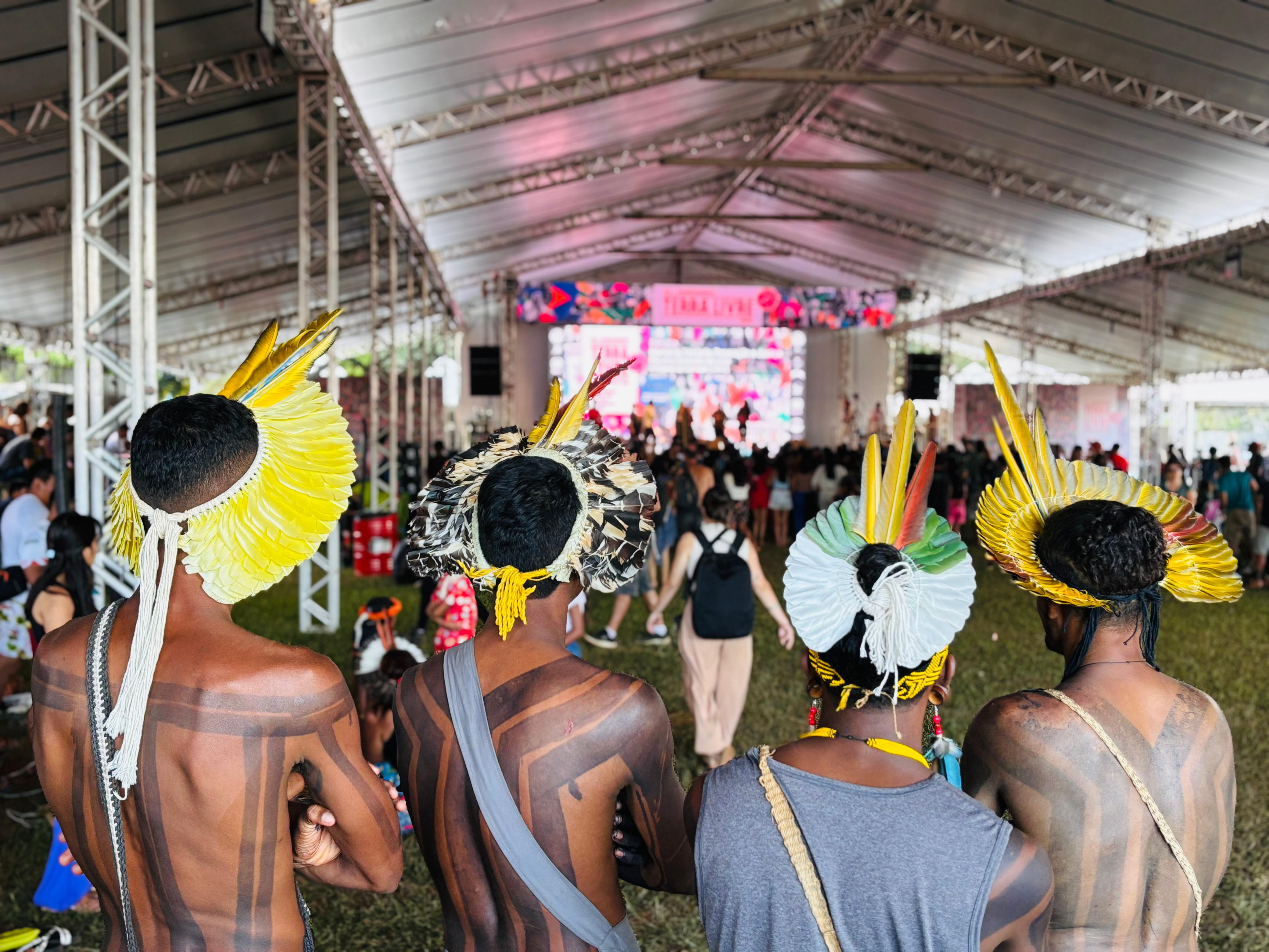 Indígenas reunidos no Acampamento Terra Livre em Brasília Um grupo de quatro indígenas com pintura corporal tradicional e cocares de penas está de costas para a câmera, voltado para um palco sob uma grande tenda. O palco está bem iluminado e apresenta artistas, enquanto uma plateia assiste.