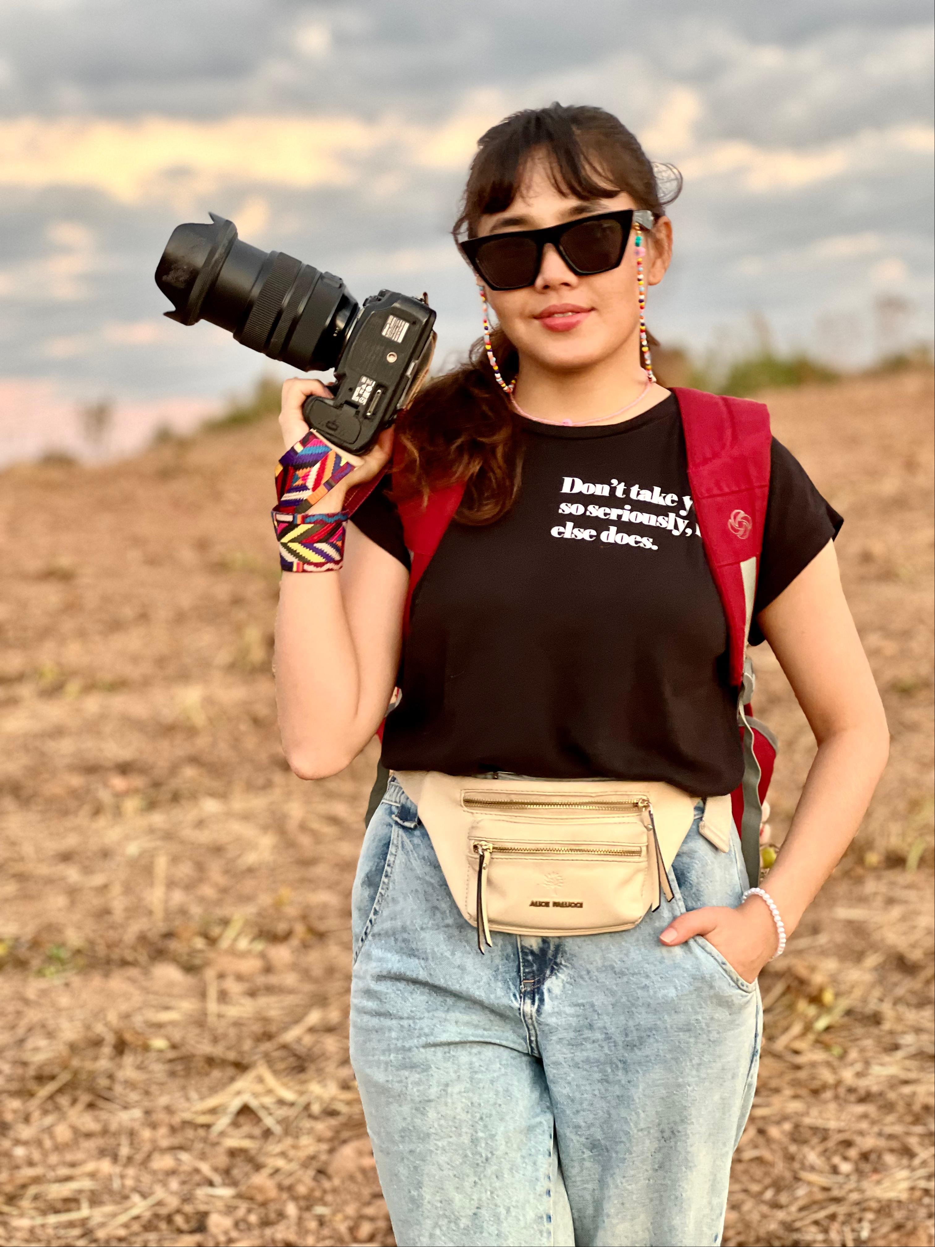 Uma mulher está de pé ao ar livre em um campo, segurando uma câmera com uma lente grande na mão direita. Ela está vestindo uma camiseta preta com texto branco. A pessoa também está usando jeans azul claro, uma pochete bege na cintura e uma mochila vermelha. O fundo mostra um campo aberto com alguma vegetação e um céu nublado.