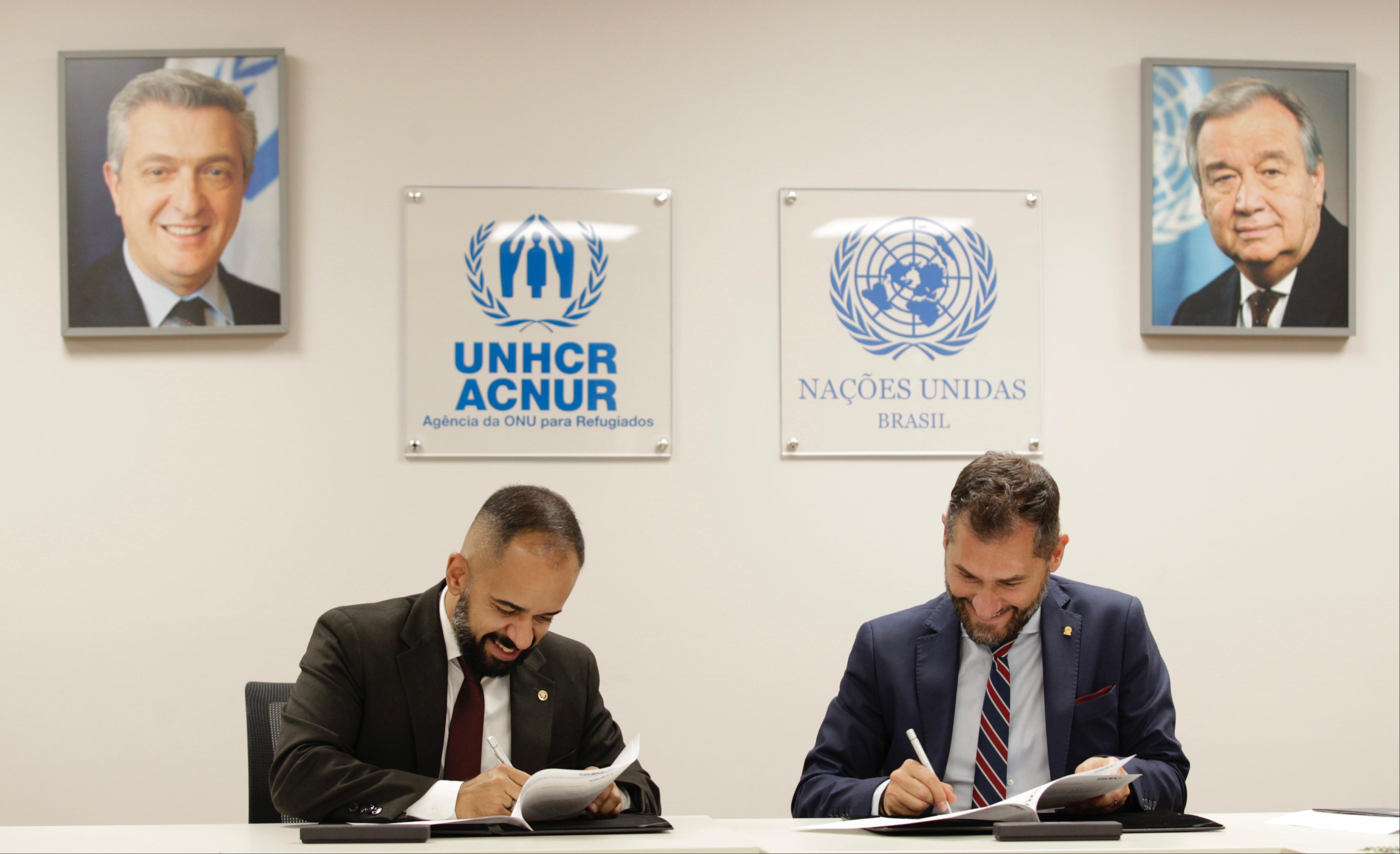 ACNUR e DPU renovam parceria Dois homens brancos, sentados em uma mesa, assinam documentos no escritório do ACNUR Brasil