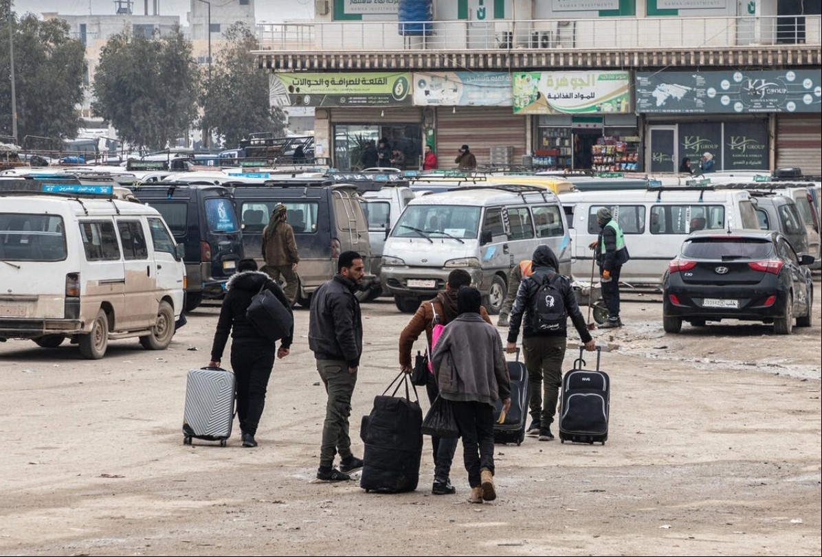 Refugiados sírios retornando da Turquia no ponto de agem da fronteira de Bab Al-Hawa Rua com várias pessoas caminhando e carregando bagagens. Há vários veículos, incluindo vans e carros, estacionados ao fundo. O cenário parece ser uma área urbana com edifícios e lojas visíveis atrás dos veículos. O chão parece molhado, possivelmente devido a uma chuva recente.
