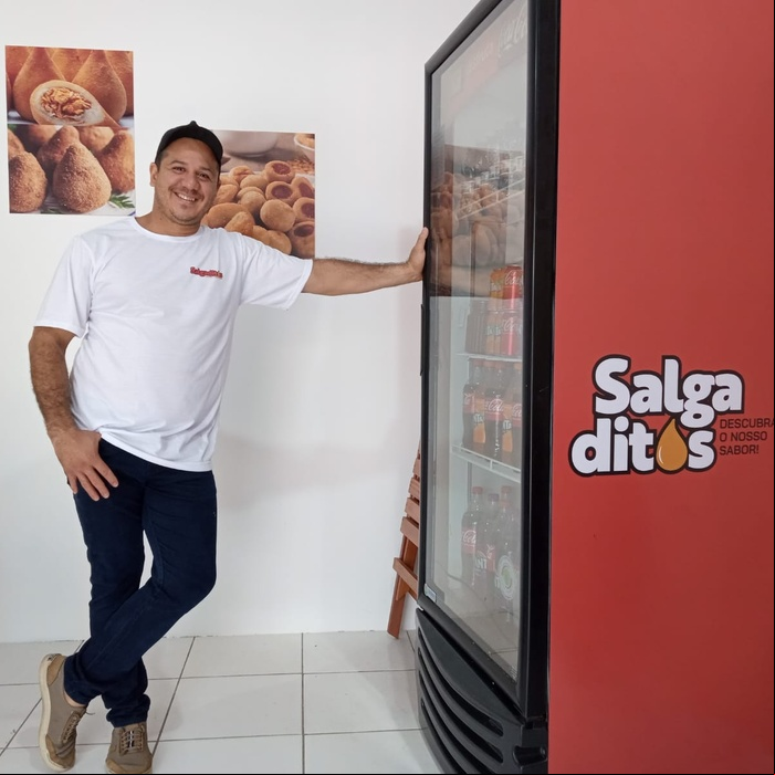 Salgaditos Um homem vestindo uma camiseta branca e calças escuras está de pé com uma mão apoiada em uma máquina de venda automática vermelha. A máquina tem o texto "Salgaditos: Descubra o Melhor Sabor" escrito nela. Há dois cartazes na parede atrás dele, ambos exibindo imagens de salgados. O chão é de azulejos e há uma porta fechada ao fundo.