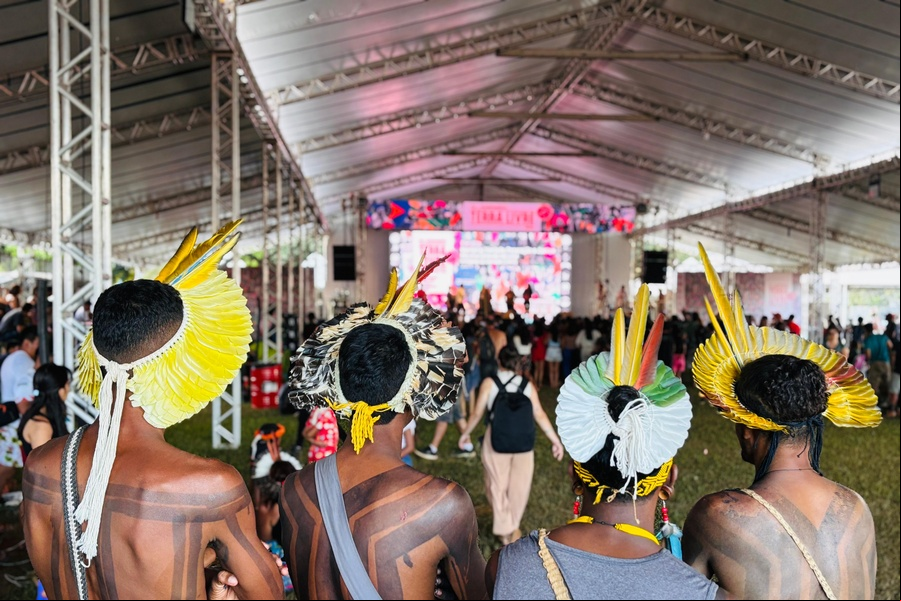 Indígenas reunidos no Acampamento Terra Livre em Brasília Um grupo de quatro indígenas com pintura corporal tradicional e cocares de penas está de costas para a câmera, voltado para um palco sob uma grande tenda. O palco está bem iluminado e apresenta artistas, enquanto uma plateia assiste.