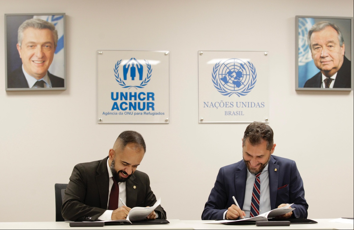 ACNUR e DPU renovam parceria Dois homens brancos, sentados em uma mesa, assinam documentos no escritório do ACNUR Brasil