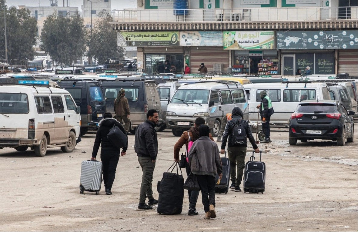 Refugiados sírios retornando da Turquia no ponto de agem da fronteira de Bab Al-Hawa Rua com várias pessoas caminhando e carregando bagagens. Há vários veículos, incluindo vans e carros, estacionados ao fundo. O cenário parece ser uma área urbana com edifícios e lojas visíveis atrás dos veículos. O chão parece molhado, possivelmente devido a uma chuva recente.