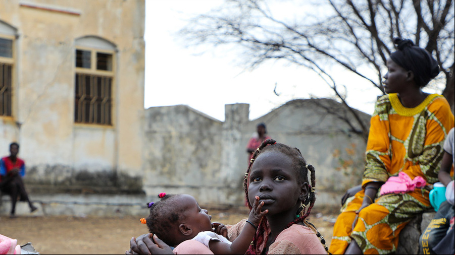 Menina sudanese no chão, com uma mala, segura uma bebê no colo, ao fundo há uma mulher