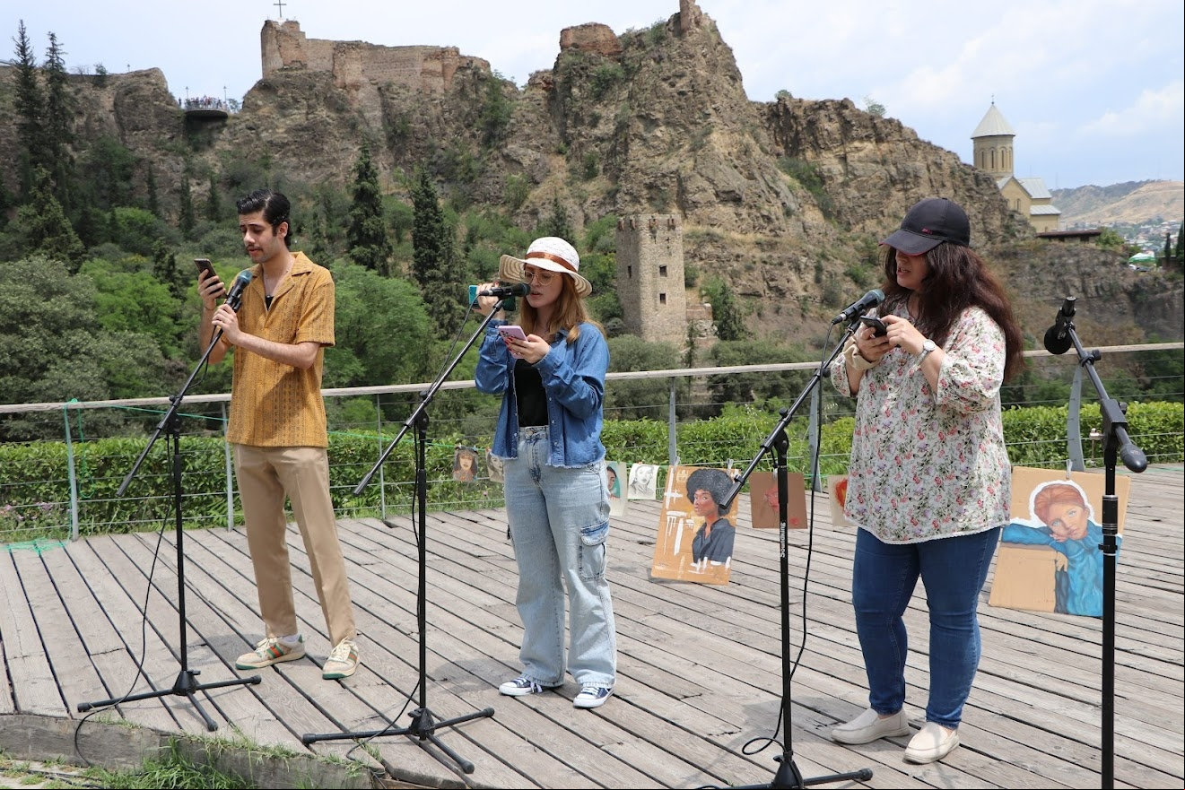 Three performers sing on an outdoor stage backed by cliffs.