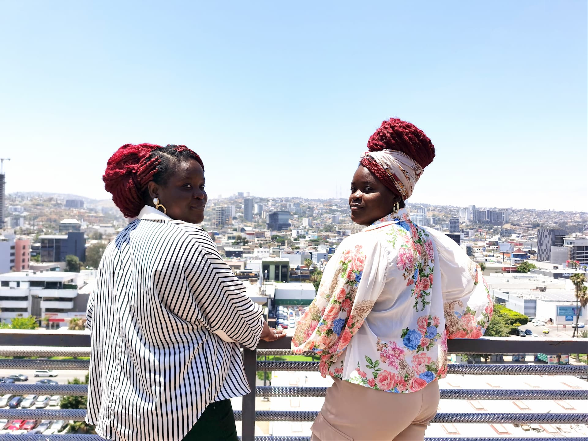 Dos mujeres miran a cámara desde un balcón en un entorno urbano.