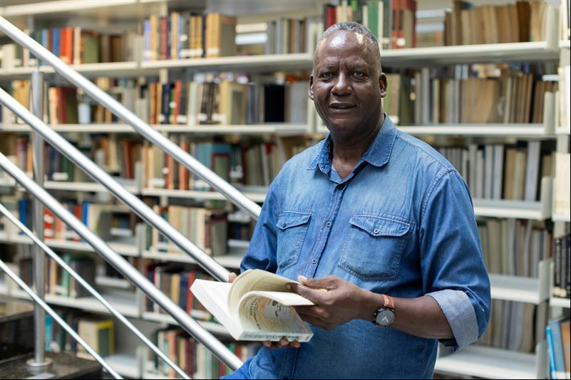 Un hombre de camisa de mezclilla posa para la foto mientras sostiene un libro en sus manos. Al fondo, libros apilados en estantes