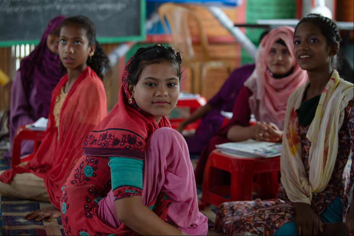 Chicas rohingyas sentadas en el piso. Algunas de ellas miran a alguien o algo fuera de cámara; una de ellas, en el centro de la imagen, ve directo a la cámara.