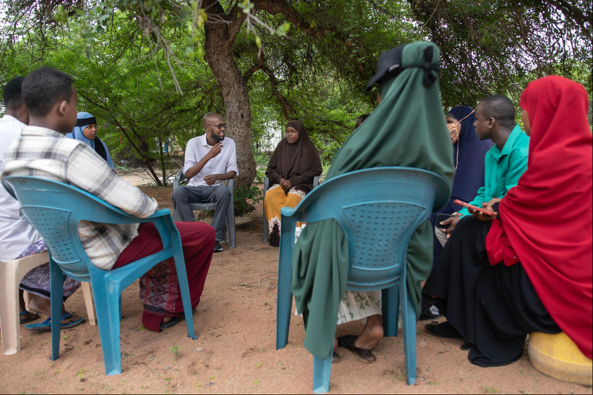 Un grupo de personas discute a fondo al aire libre, bajo los árboles, en sillas de plástico.