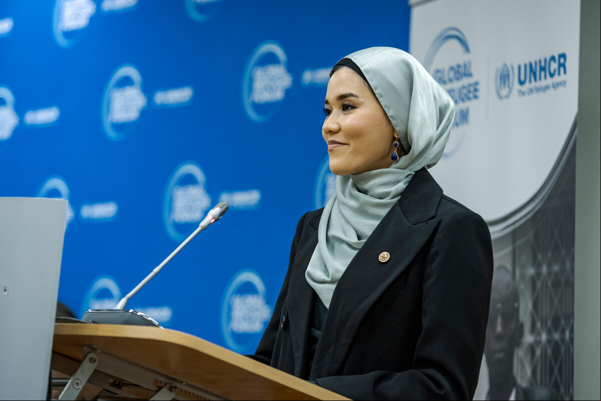Una mujer, que lleva puesto un hijab, pronuncia un discurso desde un podio. Frente a ella, un delgado micrófono; detrás, un fondo azul con el logotipo del Foro Mundial sobre los Refugiados.,