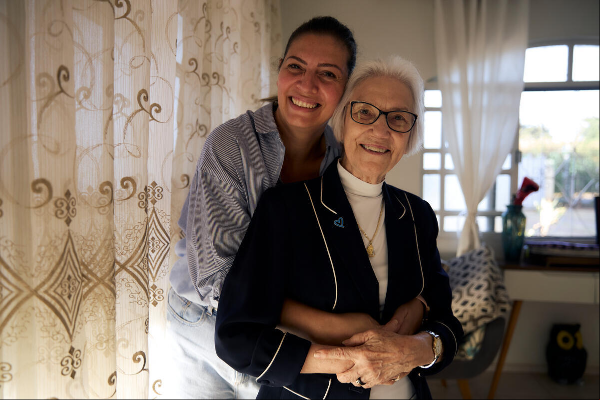 Una mujer sonriente está de pie detrás de otra que la rodea con los brazos junto a una ventana con cortinas en el interior de una casa