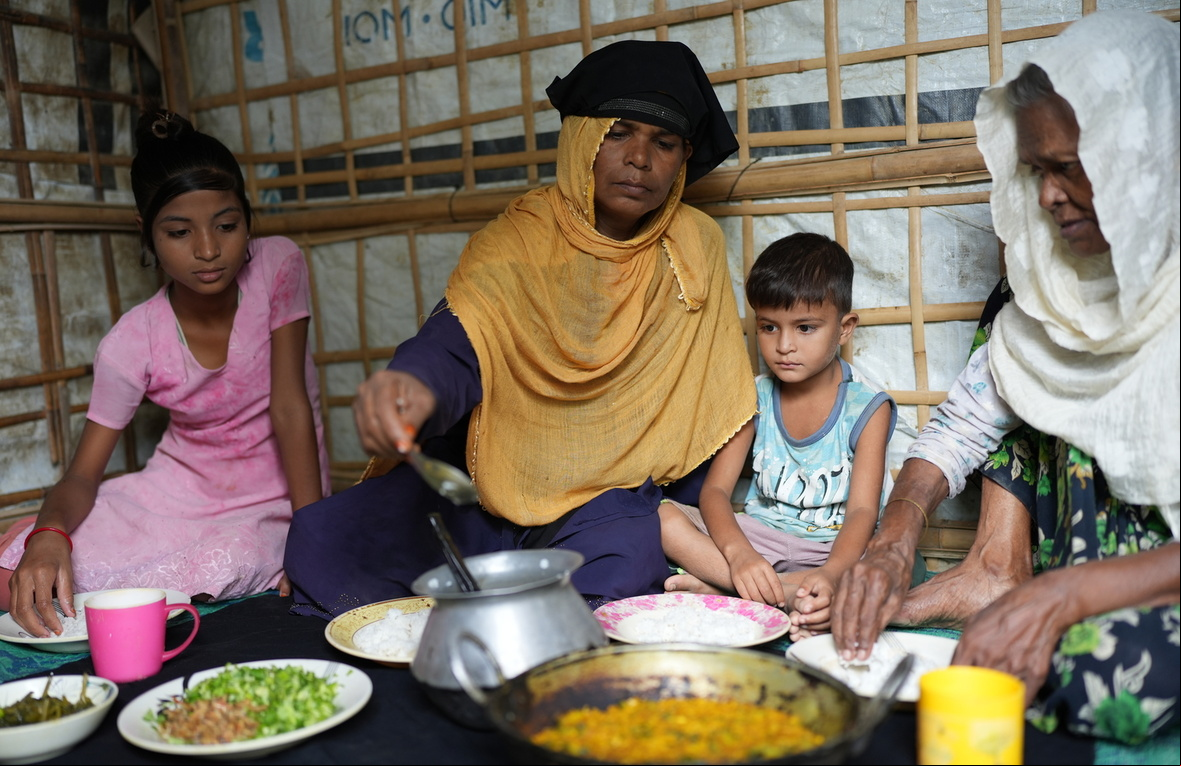 A mother and a grandmother eat a meal sitting on the floor of their shelter with two young children. Una madre y una abuela comen sentadas en el suelo de su alojamiento con dos niños pequeños.