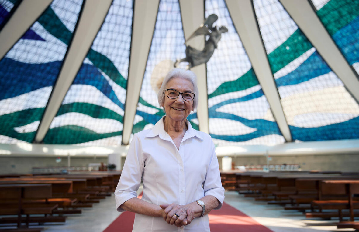 Una mujer de cabello blanco con lentes y camisa blanca en el interior de una gran iglesia con cúpula de cristal