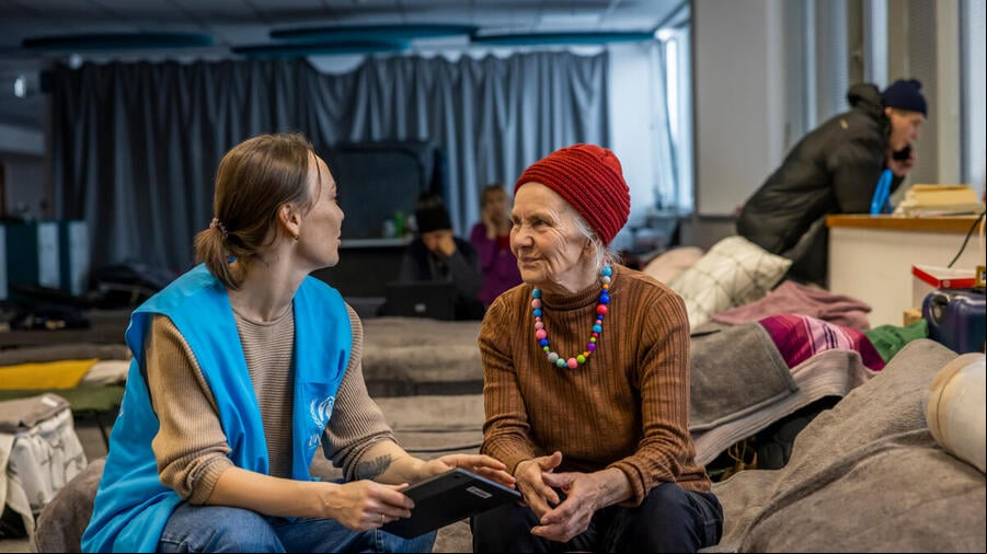 Una trabajadora de ACNUR charla con una mujer mayor; ambas están sentadas en una cama en un albergue.