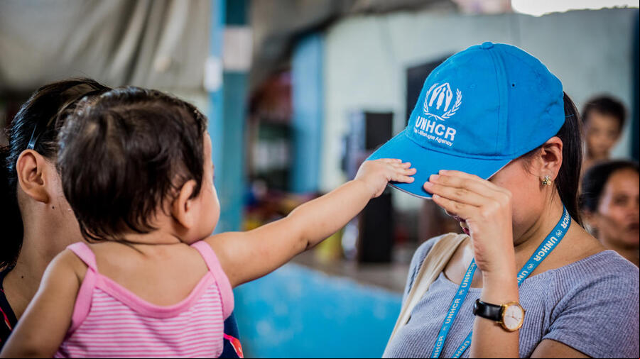 Una niña pequeña toma la gorra azul de ACNUR que lleva puesta una mujer. Una niña pequeña toma la gorra azul de ACNUR que lleva puesta una mujer.