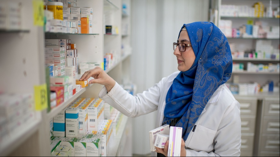 Una mujer joven, que usa anteojos y lleva puesto un hiyab azul y una bata de laboratorio, acomoda medicamentos en un estante