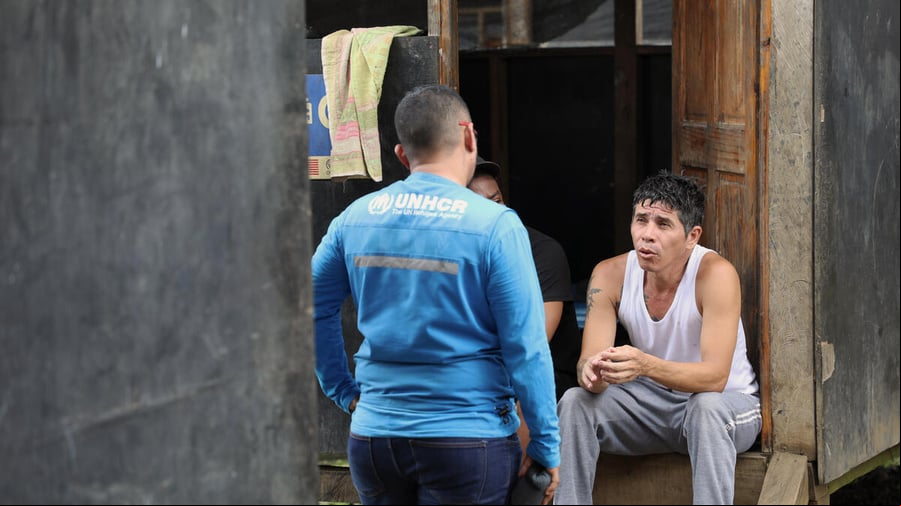 Sentado frente a una puerta de madera, un hombre joven charla con otro que está de pie delante suyo
