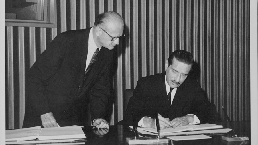 Imagen en blanco y negro. Dos hombres (uno, sentado; el otro, de pie a su lado). El hombre sentado, que aparece a la derecha, está firmando un documento; el otro observa.