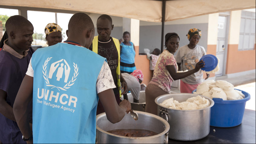 Un grupo de personas se dispone a repartir alimentos. Frente a ellas, un trabajador de ACNUR que lleva puesto un chaleco azul con el logotipo de la organización.