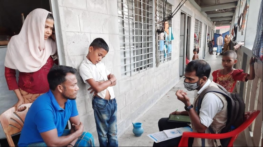 Un trabajador humanitario charla con una familia: el padre está sentado en una silla en la puerta del hogar; detrás suyo, la madre, quien lleva puesto un velo.